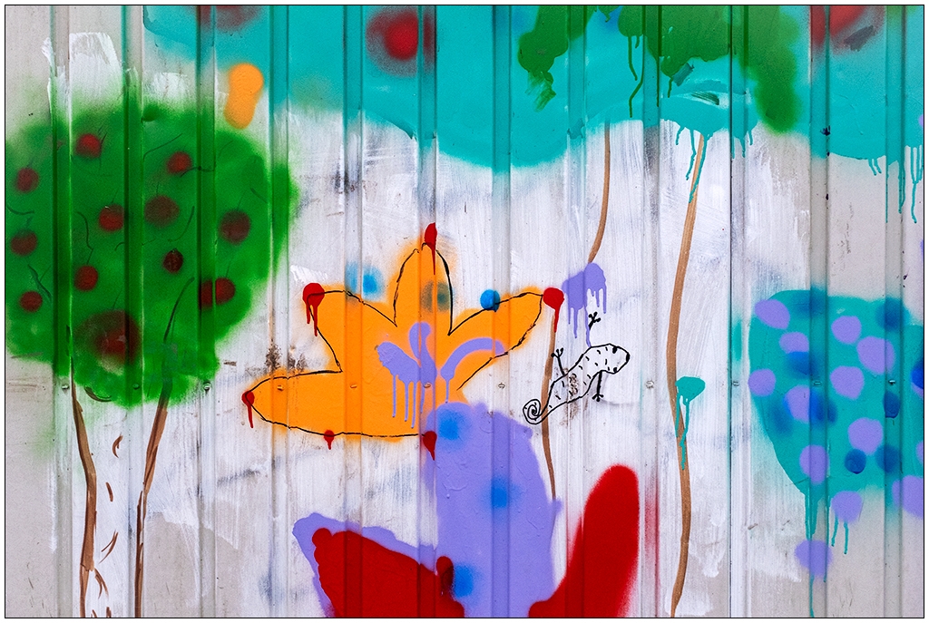 Fotografie, die eine Wand eines bemalten Bauwagens zeigt. Der Bauwagen ist bunt bemalt, wahrscheinlich von Kindern. Links ist ein Baum zu erkennen, rechts daneben eine Blüetnpflanze in Orange, davon rechts eine kleine Echse.