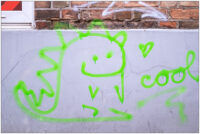 Fotografie, die eine Hauswand mit einem Graffiti zeigt. Im oberen Teil sind drei Reihen Ziegelsteine zu sehen. Der untere Teil der Hauswand ist verputzt und grau angestrichen. Auf die graue Fläche ist in leuchtend grün ein kleiner Drache gesprüht. Der Drache guckt freundlich und lächelt. Rechts neben dem Drachen ist ein kleines Herz hingesprüht und darunter das Wort "cool". Alles ist in leuchtendem Grün gesprüht.