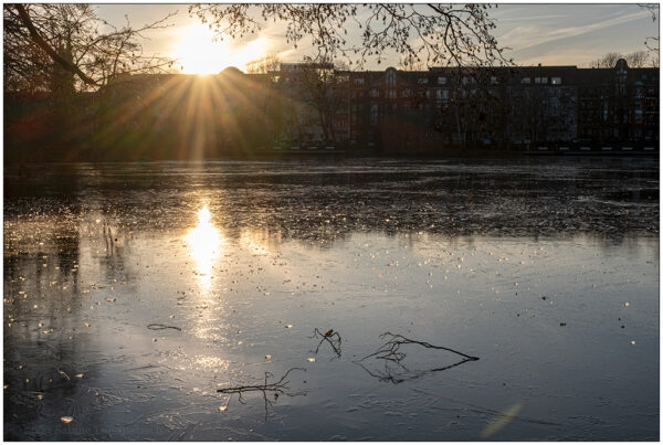 Foto mit Blick auf den zugefrorenen Schreventeich im Kieler Schrevenpark. Das Foto ist eine Gegenlichtaufnahme. Im Vordergrund liegen ein paar kleinere Äste auf dem zugefrorenen Teich.