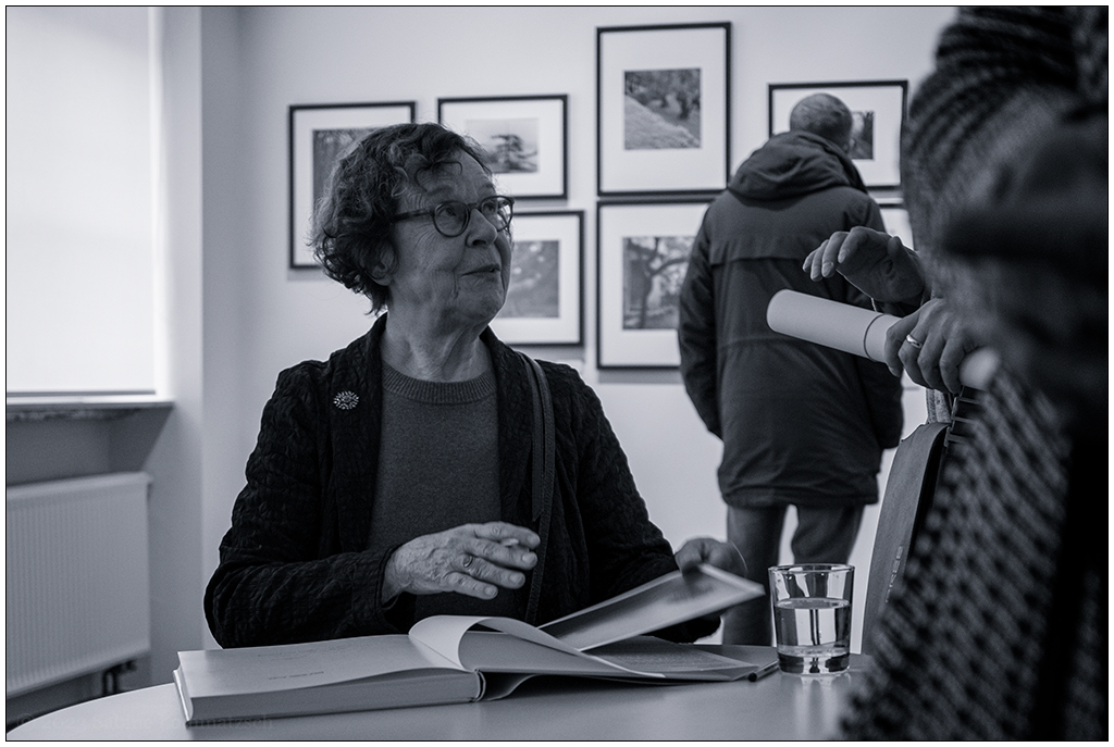 Die Fotografin Barbara Klemm sitzt an einem Tisch und schaut zu einer Person herauf, die ihr gerade ein Buch zum Signieren auf den Tisch gelegt hat. Im Hintergrund sind mehrere Fotos in einem Ausstellungsraum zu sehen. Ein Mann betrachtet ein Bild der Ausstellung.