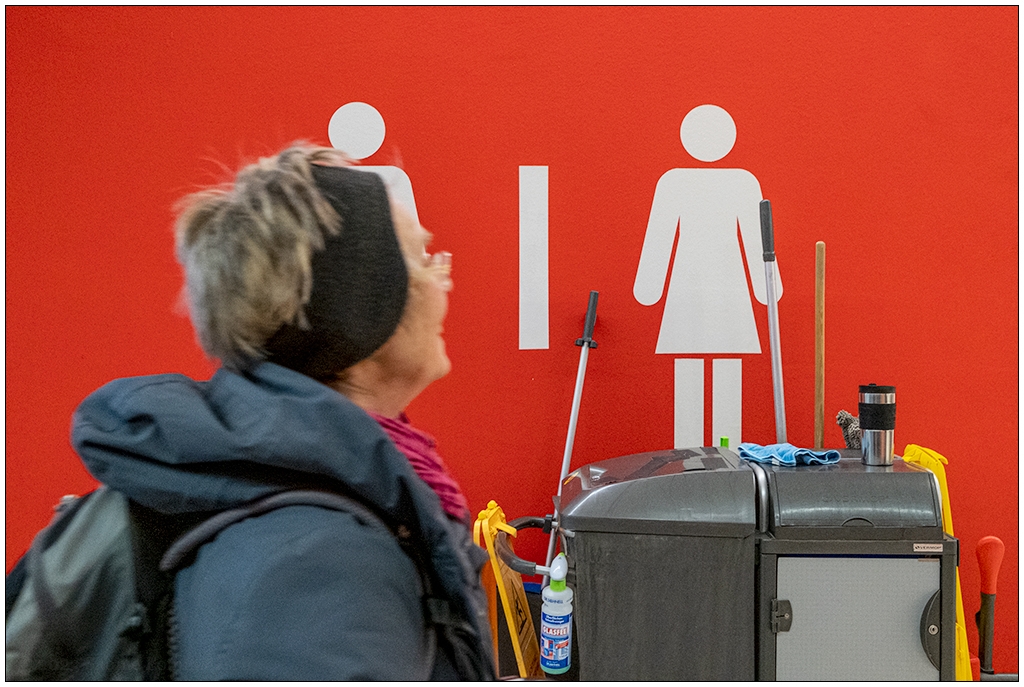 Eine rote Wand mit dem Zeichen für Mann und Frau (Toilette). Darunter ein Puztwagen. Eine Frau mit kurzen Haaren und Stirnband läuft von links ins Bild hinein.