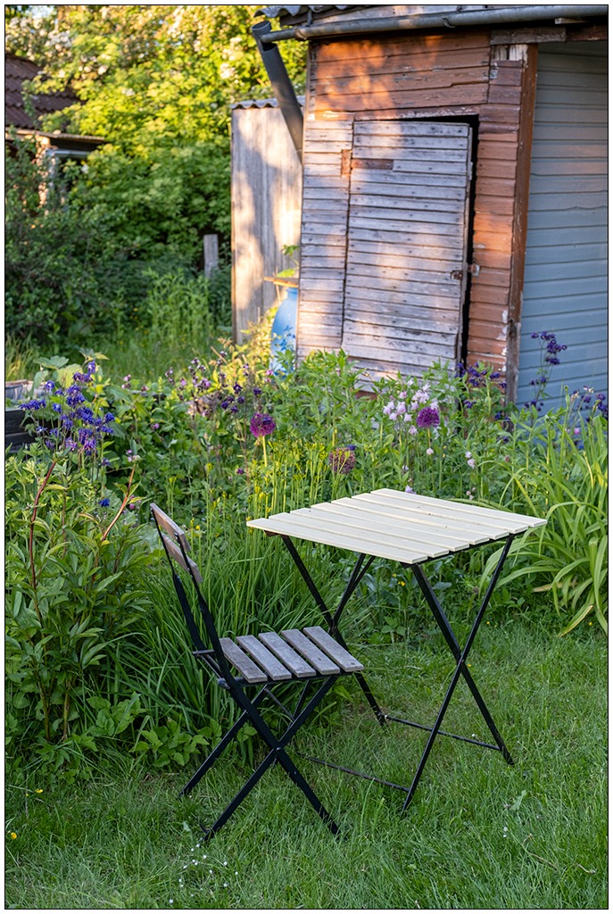Gartenidylle mit einem kleinen Gartentisch samt Stuhl. Im HIntergrund ein schiefer Holzschuppen.