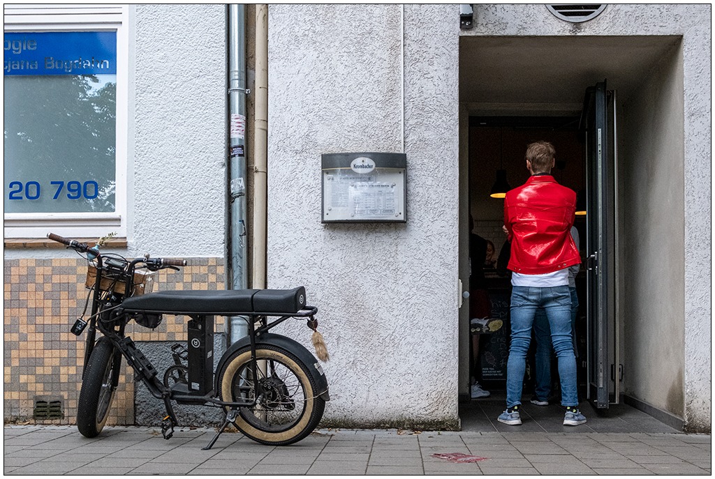 Ein Mann in blauer Jeans und mit einer roten Lederjacke steht in einem offenen Eingang zu einem Restaurant. Links neben ihm steht ein außergewöhnliches Elektromotorrad. Der Mann ist nur von hinten zu sehen.