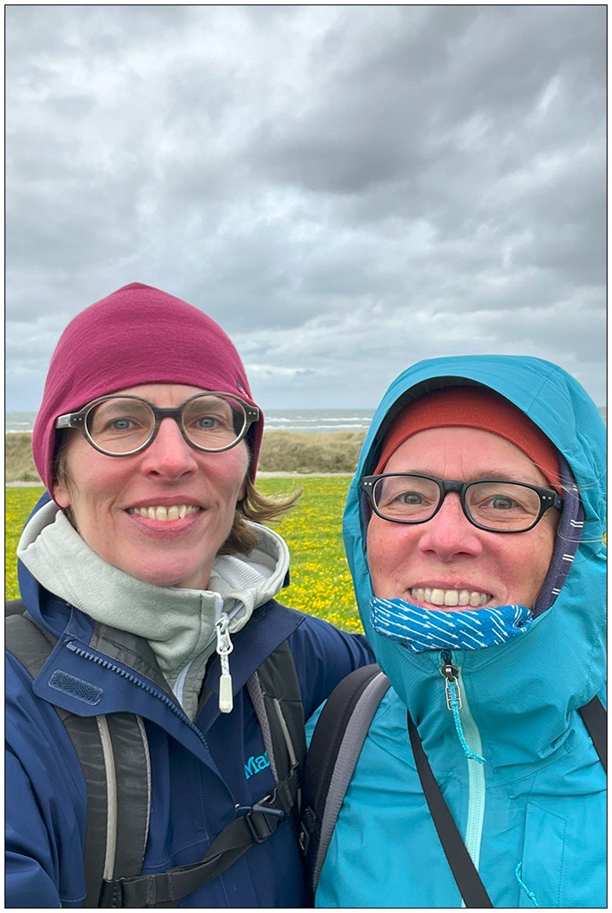 Zwei Frauen mit Mützen auf schauen in die Kamera. Im Hintergrund die durch einen Sturm aufgewühlte Ostsee. Der Himmel ist grau.