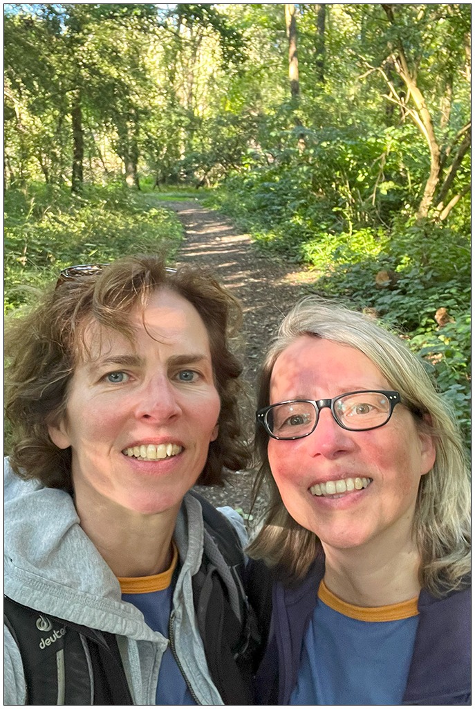 Zwei Frauen blicken in die Kamera, im Hintergrund ein Wanderweg. Die Sonne scheint.