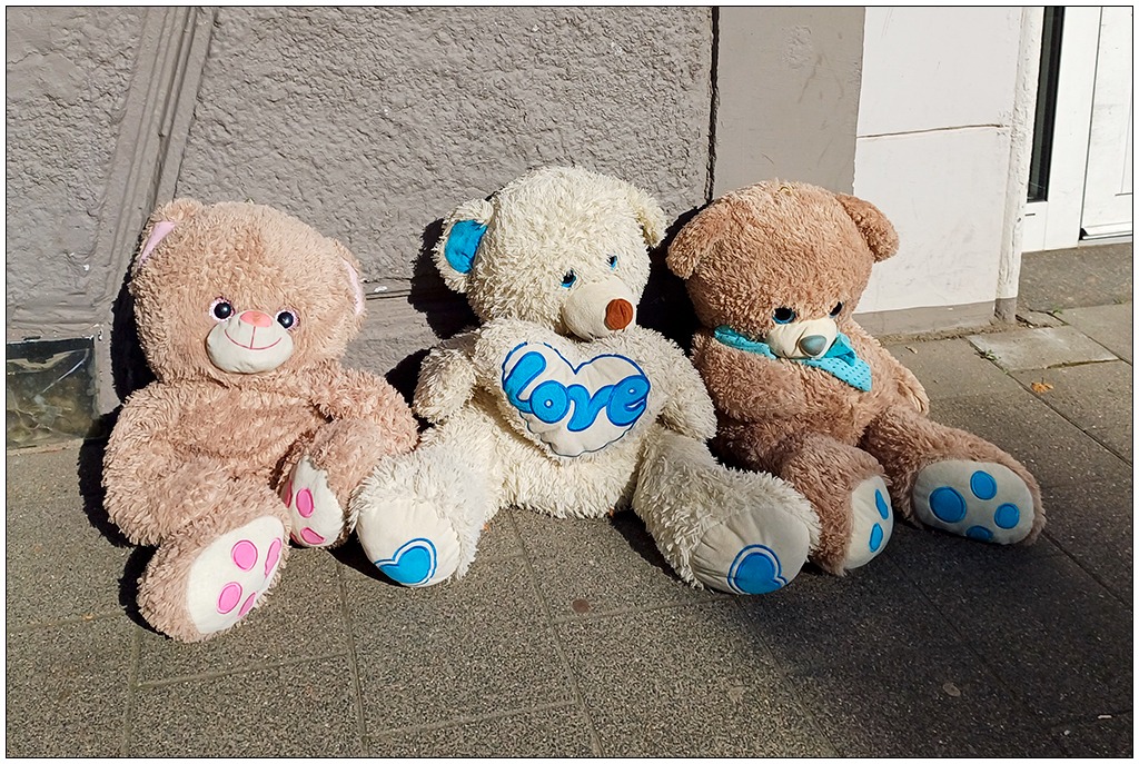 Drei Teddys liegen angelehnt an eine Hauswand auf dem Bürgersteig in der Sonne.