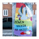 An einem Laternenpfahl in Kiel klebt ein Aufkleber mit einer Möwe. Die Möwe trägt einen Schal und eine Mütze in Regenbogenfarben. Unter der Möwe steht: Möwen kacken auf Nazis!